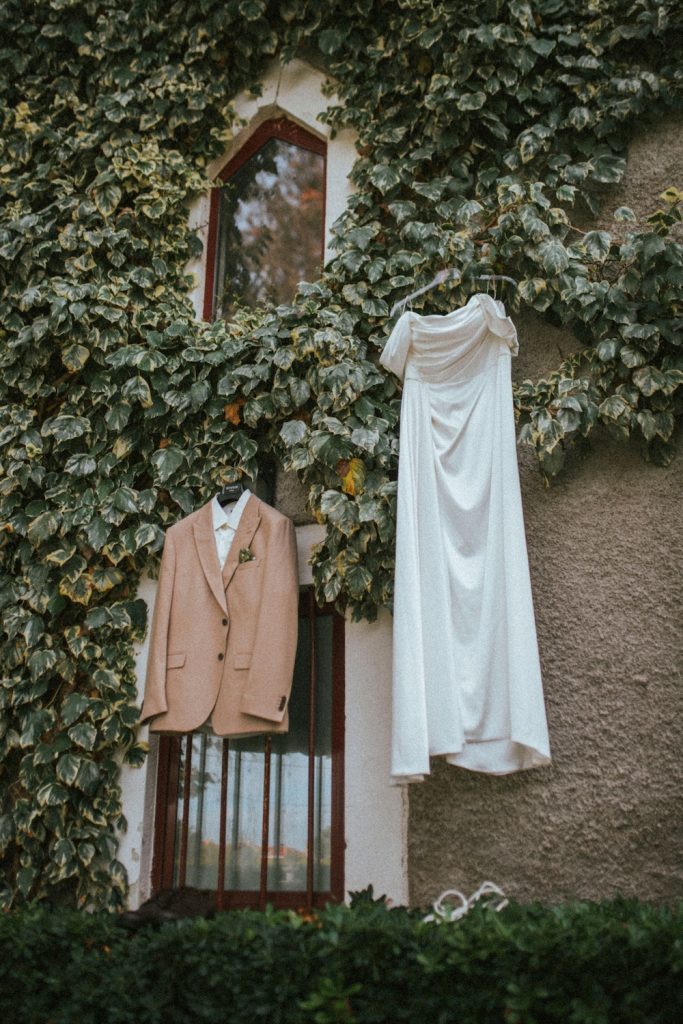 Tan suit jacket and white wedding dress hanging from the outside of a ivy-covered building